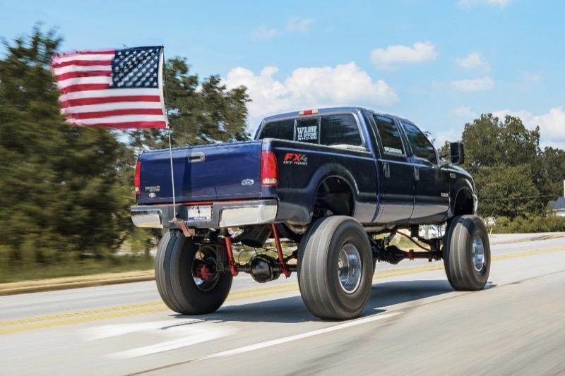 Lifted Ford F-250 on big tires driving down the highway with an American flag.